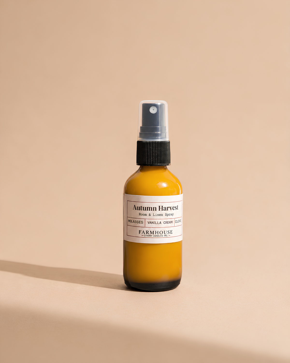 A small amber glass spray bottle with a white label reading 'Autumn Harvest' by Farmhouse Charm Candles on a warm beige background.