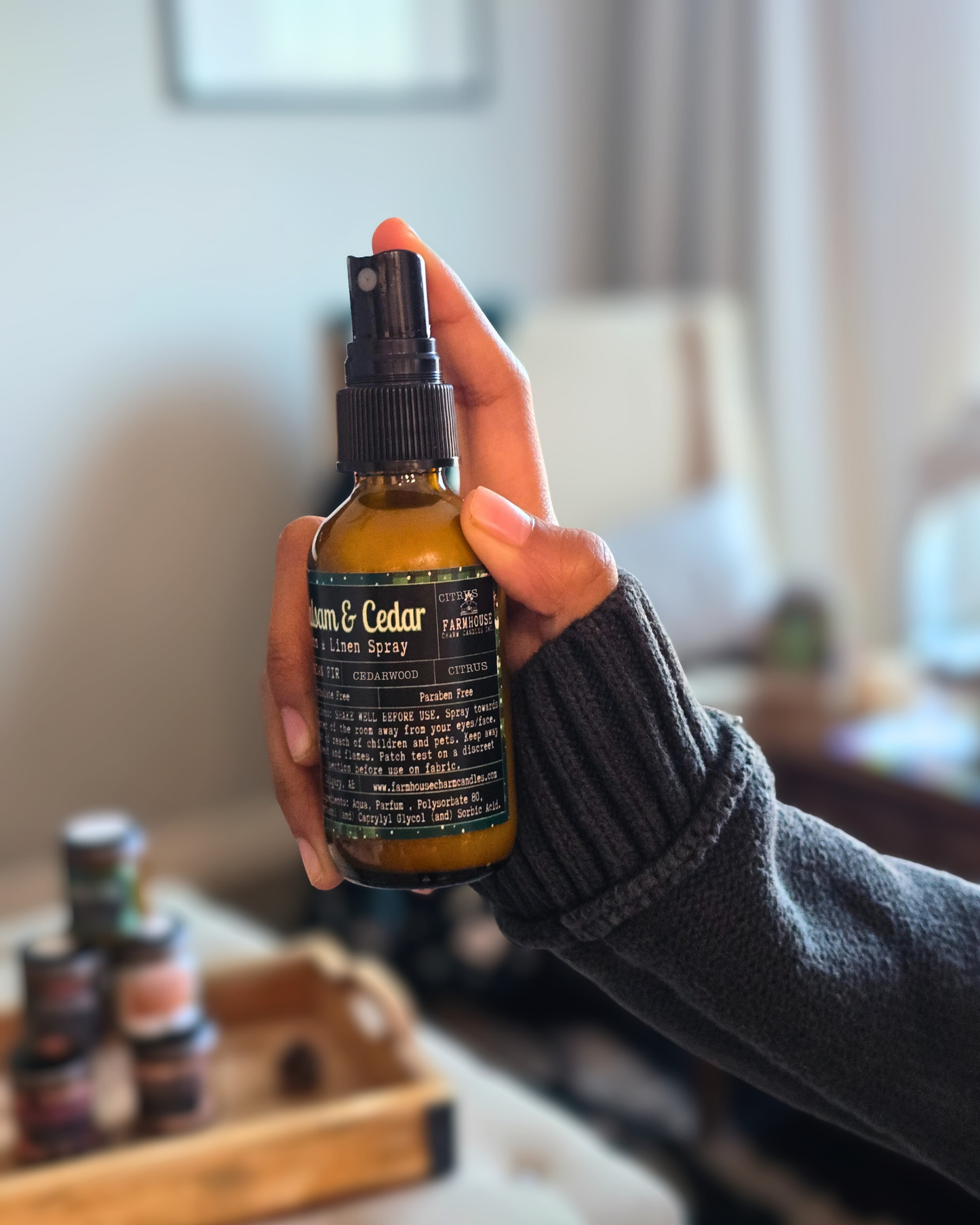 Hand holding Amber bottle of Balsam & Cedar Room & Linen Spray by Farmhouse Charm Candles, styled on a cozy winter backdrop with pine branches and berries, evoking a fresh woodsy and cabin-like scent.