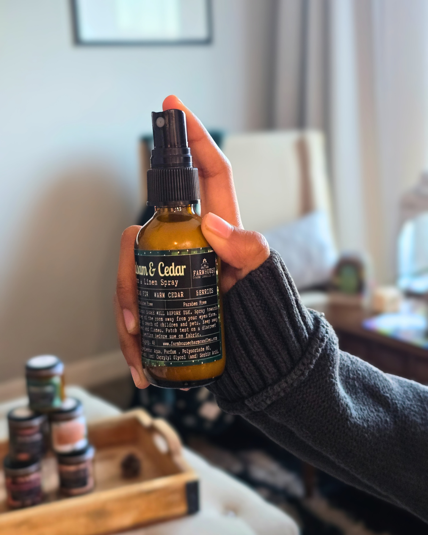 Hand holding a bottle of 'Cedar & Cedar Room & Linen Spray by Farmhouse Charm Candles with a blurred indoor background