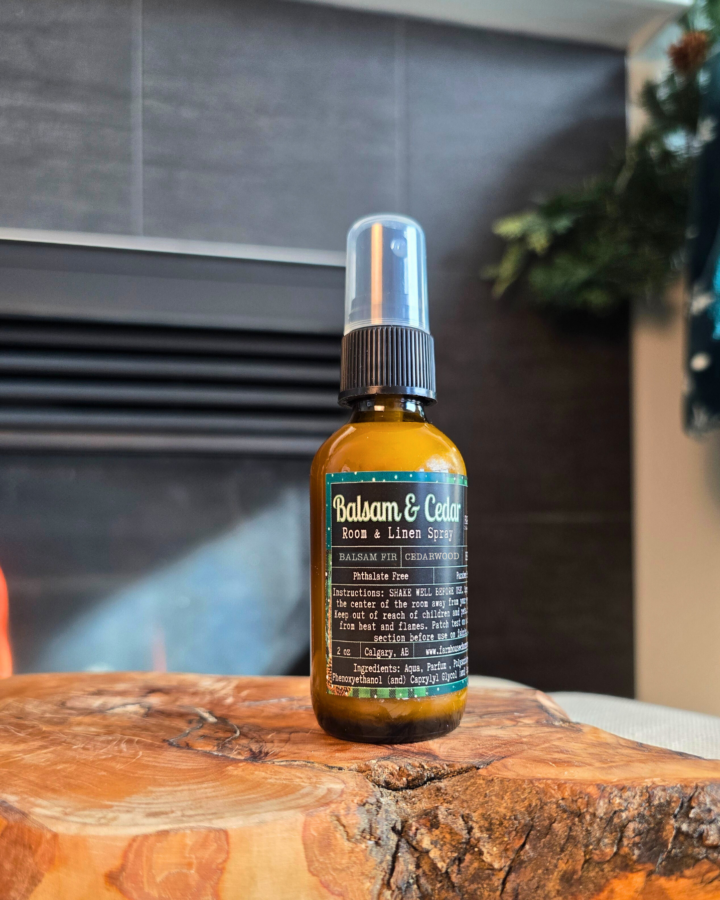 Amber bottle of Balsam & Cedar Room & Linen Spray by Farmhouse Charm Candles, styled on a cozy winter backdrop with pine branches and berries, evoking a fresh woodsy and cabin-like scent.