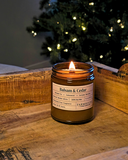 A lit amber glass jar candle labeled 'Balsam & Cedar' by Farmhouse Charm Candles., placed on a rustic wooden surface with a blurred Christmas tree and warm lights in the background.