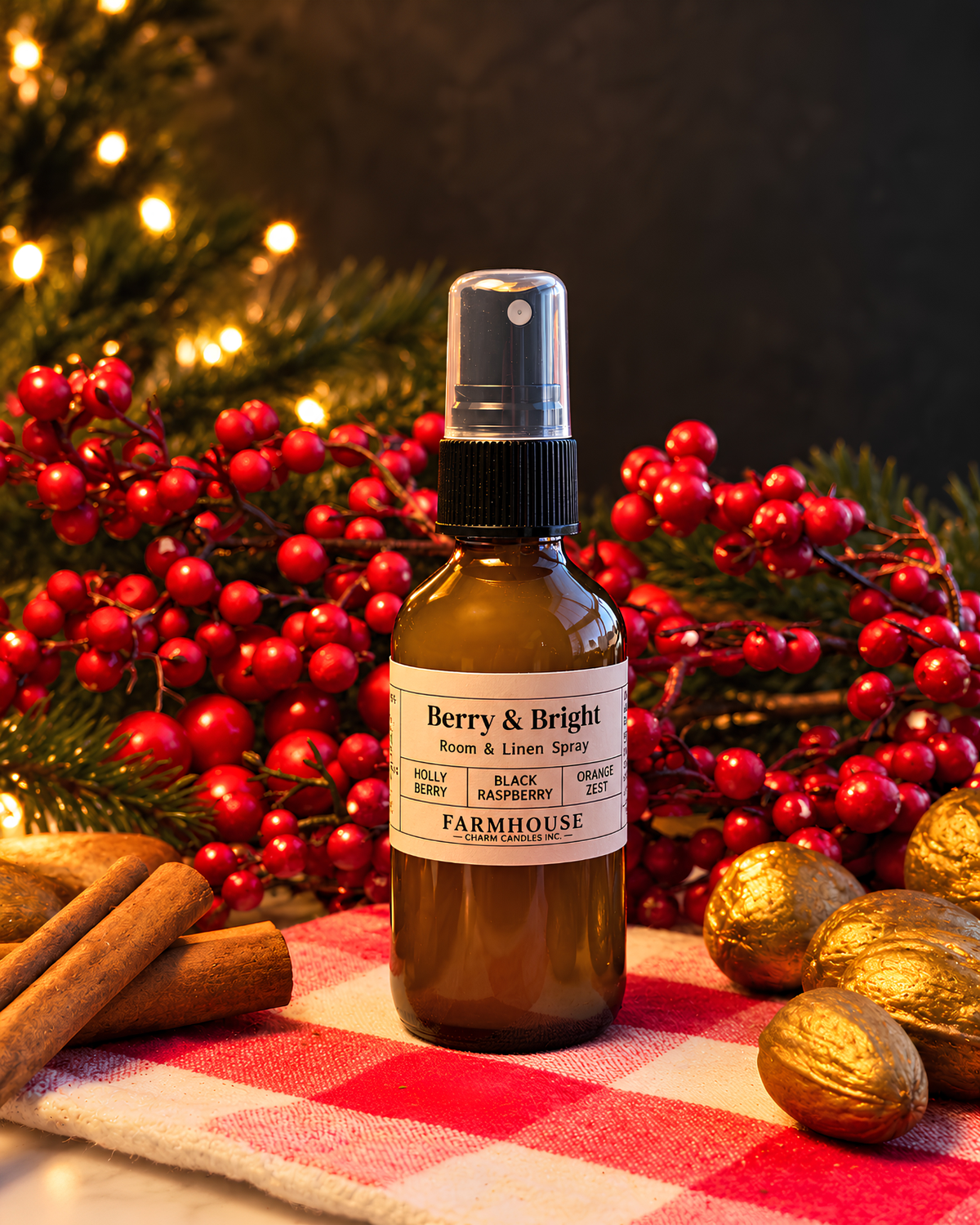 A brown glass room and linen spray bottle with a black spray top and a white label reading 'Berry & Bright' by 'FARMHOUSE Charm candles styled on a red-and-white checkered cloth with cinnamon sticks, gold pinecones, and red berries against a dark background with blurred holiday lights.