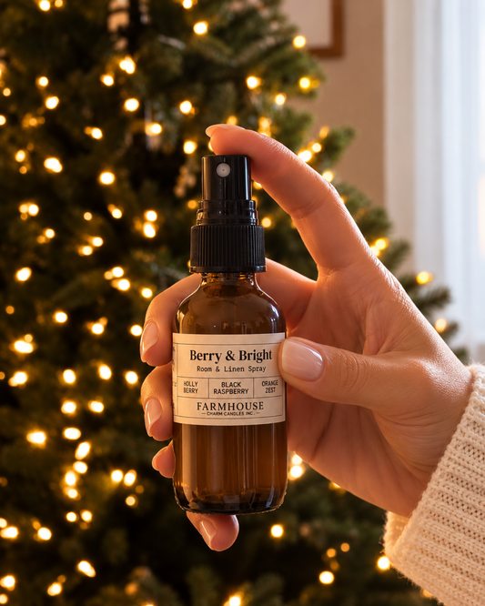 A hand holds a small amber glass spray bottle labeled Berry & Bright Room & Liquid Spray by FARMHOUSE Charm candles, with a blurred Christmas tree and warm lights in the background.