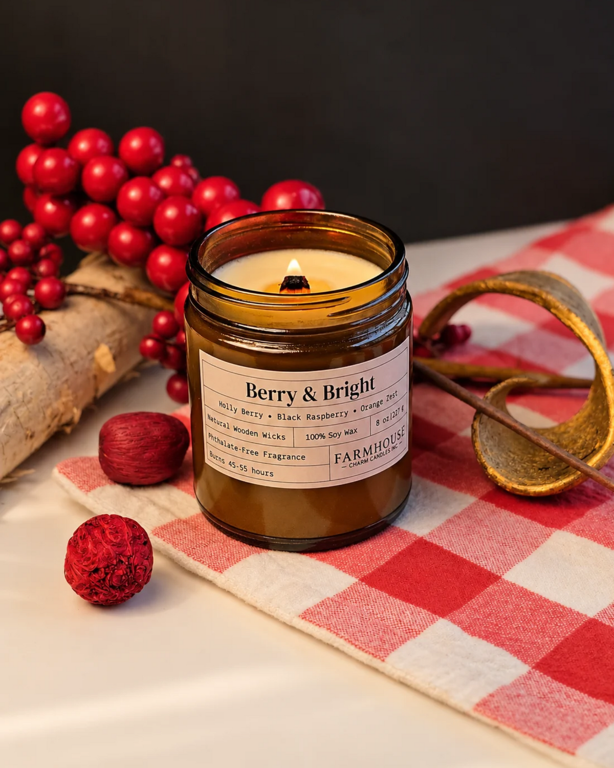 An amber glass jar candle with a lit wick sits on a red-and-white checkered cloth, surrounded by red berries, a log slice, and a gold-toned decorative object. The front label reads Berry & Bright by Farmhouse Charm Candles
