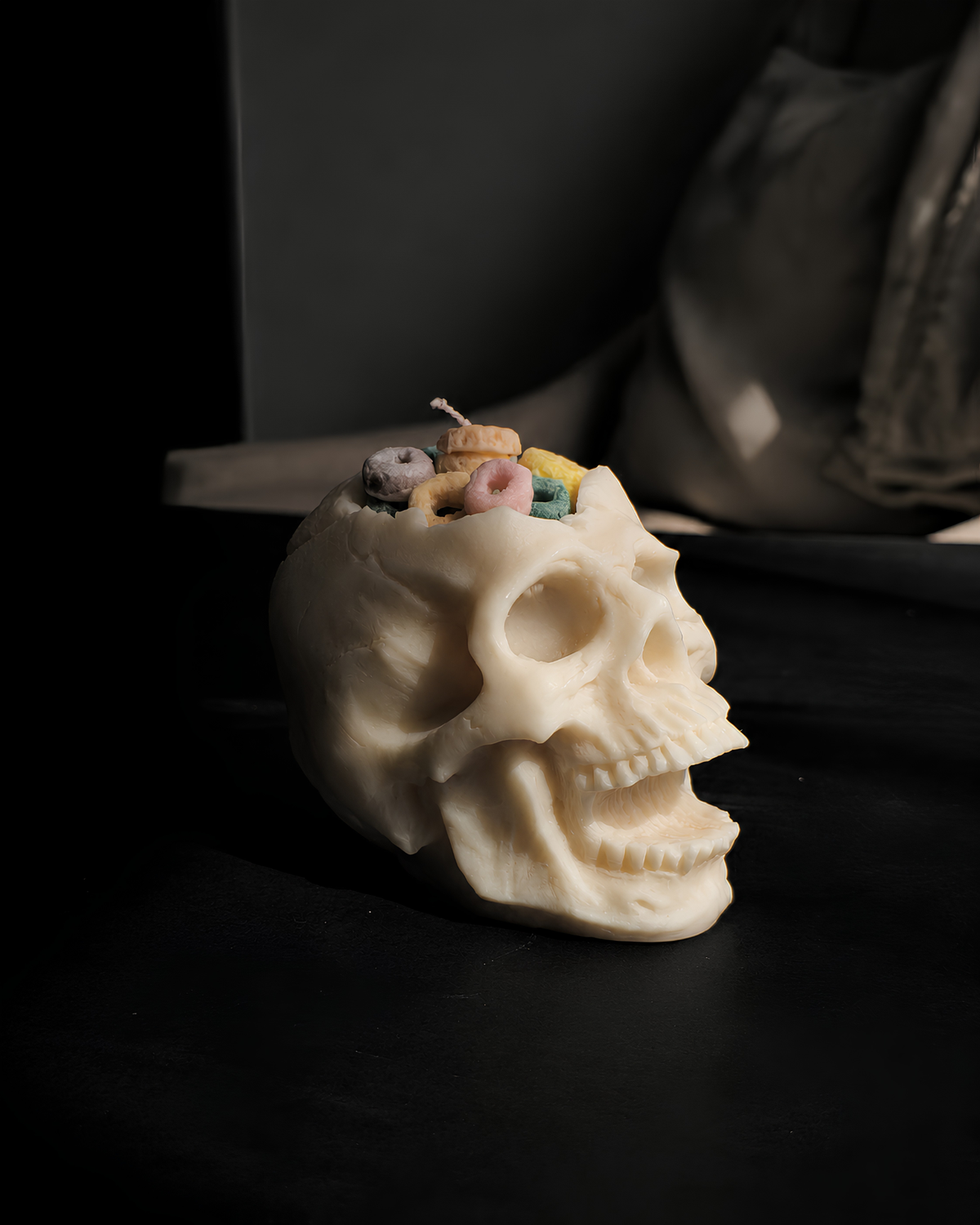 A cream-colored skull-shaped candle with colorful candy-like details inside the cranium, set on a dark surface with a moody, dimly lit background. Cereal Killer Pillar Candle by Farmhouse Charm Candles