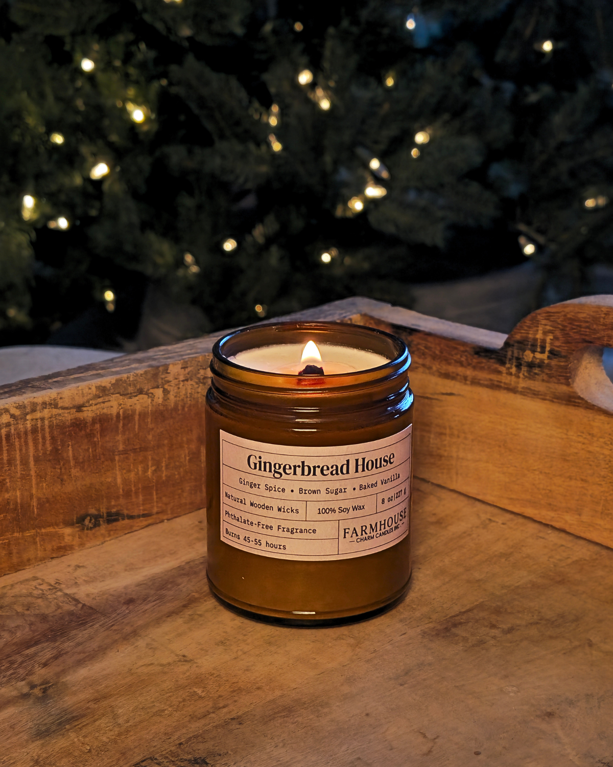 A lit amber glass jar candle labeled 'Gingerbread House' by Farmhouse Charm Candles placed on a rustic wooden surface with a blurred Christmas tree and warm string lights in the background.