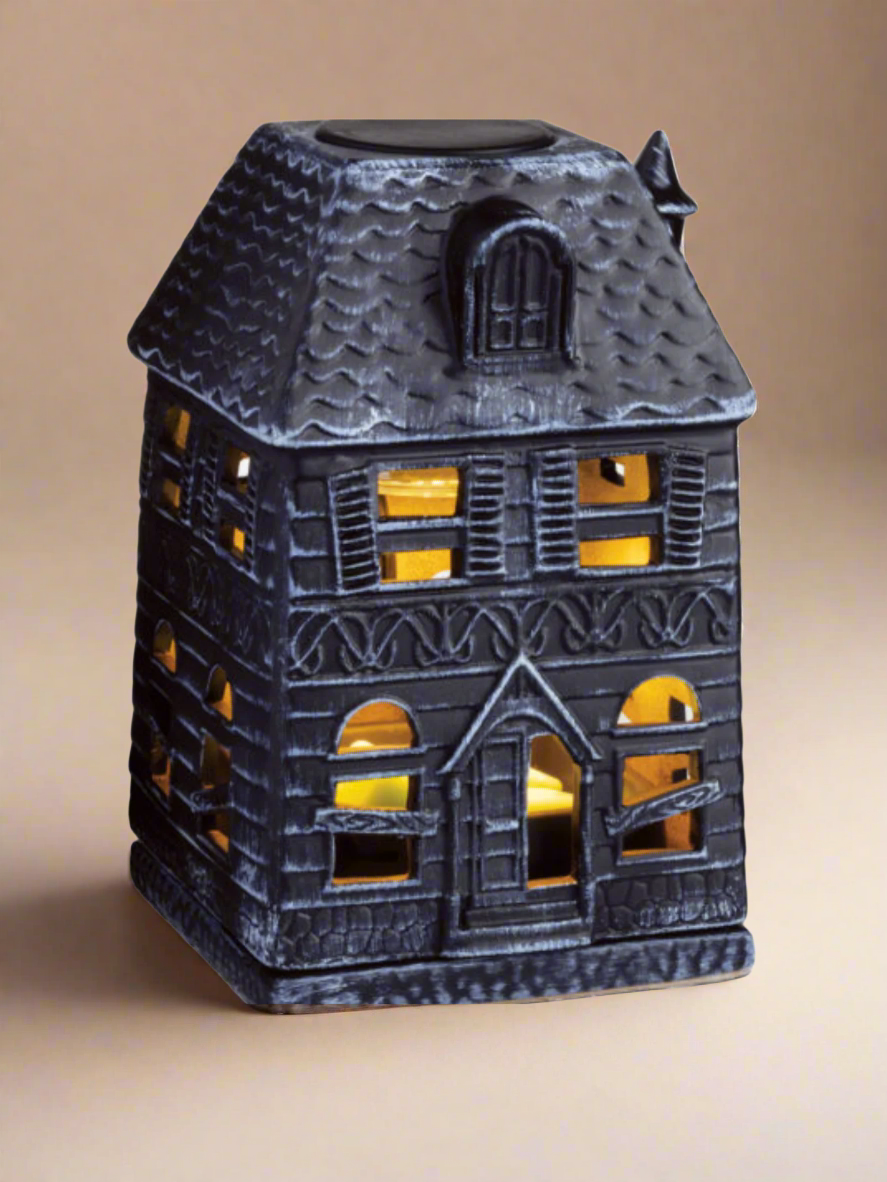 Porcelain Haunted House Illumination Fragrance Warmer with boarded-up windows casting a soft glow. Designed to safely melt wax melts and fill the home with autumn scents. Features a spooky haunted mansion design, includes a warming bulb, and operates with a 3 ft cord and on/off switch. Perfect for Halloween and fall décor. www.farmhousecharmcandles.com