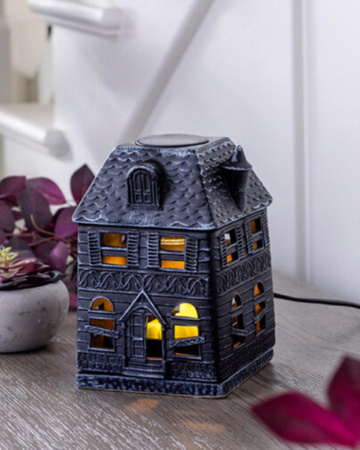 Porcelain Haunted House Illumination Fragrance Warmer with boarded-up windows casting a soft glow. Designed to safely melt wax melts and fill the home with autumn scents. Features a spooky haunted mansion design, includes a warming bulb, and operates with a 3 ft cord and on/off switch. Perfect for Halloween and fall décor. www.farmhousecharmcandles.com