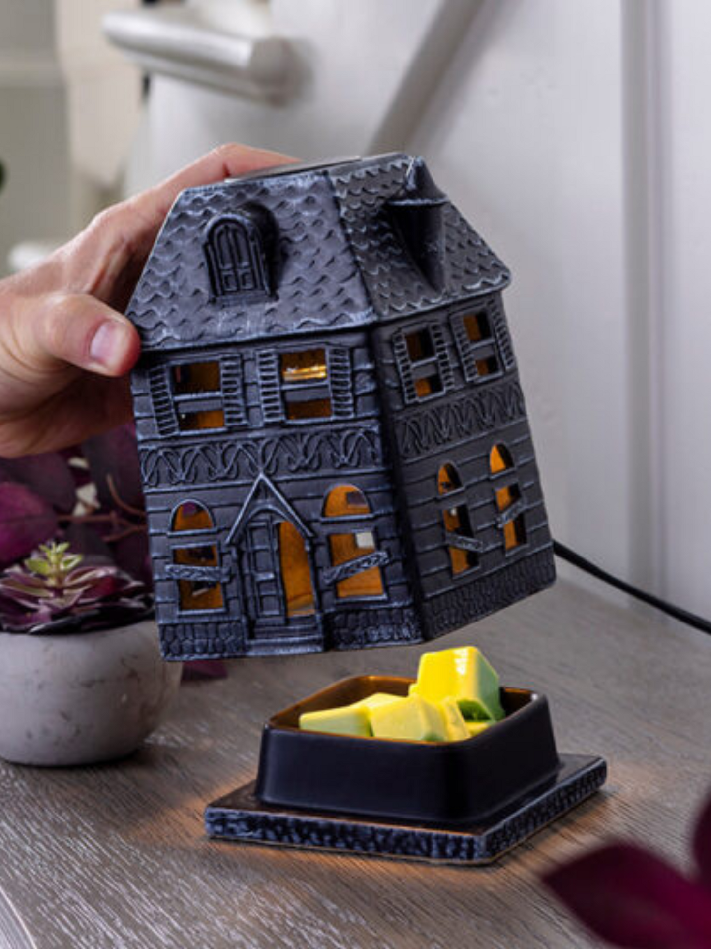 Porcelain Haunted House Illumination Fragrance Warmer with boarded-up windows casting a soft glow. Designed to safely melt wax melts and fill the home with autumn scents. Features a spooky haunted mansion design, includes a warming bulb, and operates with a 3 ft cord and on/off switch. Perfect for Halloween and fall décor. www.farmhousecharmcandles.com
