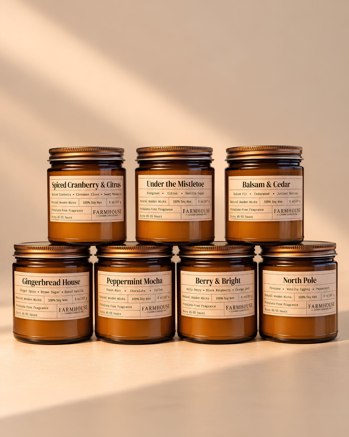 7 amber glass jar candles with gold lids and cream labels arranged in two rows on a warm beige surface. The labels show different scent names including Spiced Cranberry & Citrus, Under the Mistletoe, Balsam & Cedar, Gingerbread House, Peppermint Mocha, Berry & Bright, and North Pole. Home for Christmas Candle Bundle by Farmhouse Charm Candles