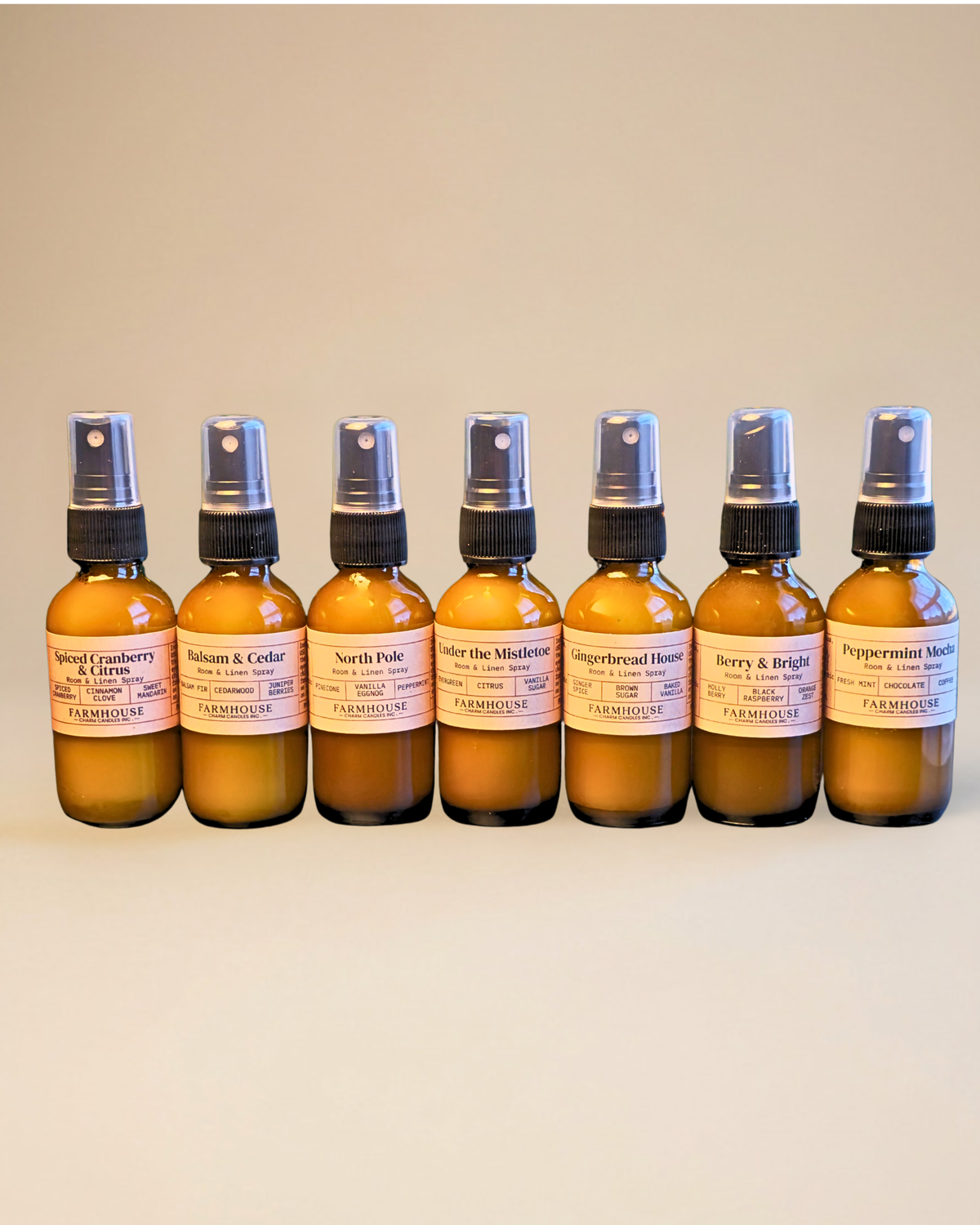 Seven amber glass spray bottles with black spray tops and white labels, arranged in a row against a plain beige background. The labels show different scent names including Balsam & Cedar, Peppermint Mocha. North Pole. Home For Christmas Room & Linen Spray by Farmhouse Charm Candles