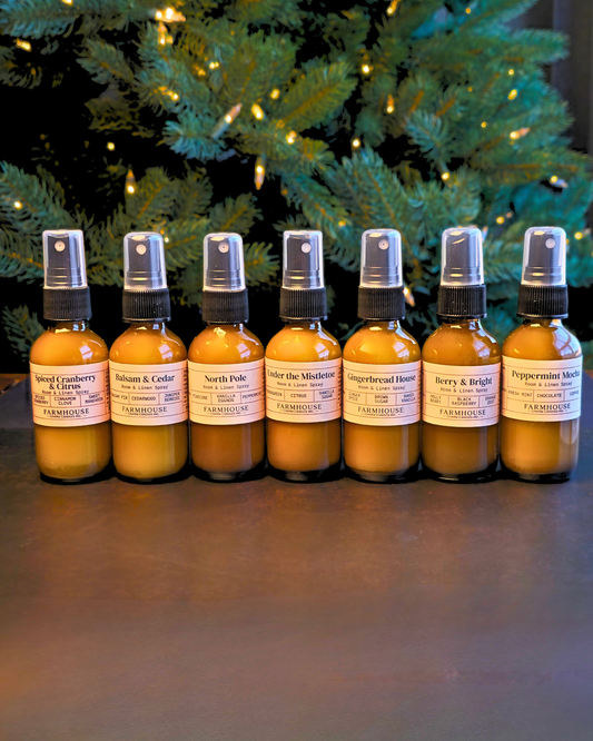 Seven amber glass spray bottles with white labels arranged in a row on a dark surface, with a blurred Christmas tree in the background. Home For Christmas Room & Linen Spray by Farmhouse Charm Candles
