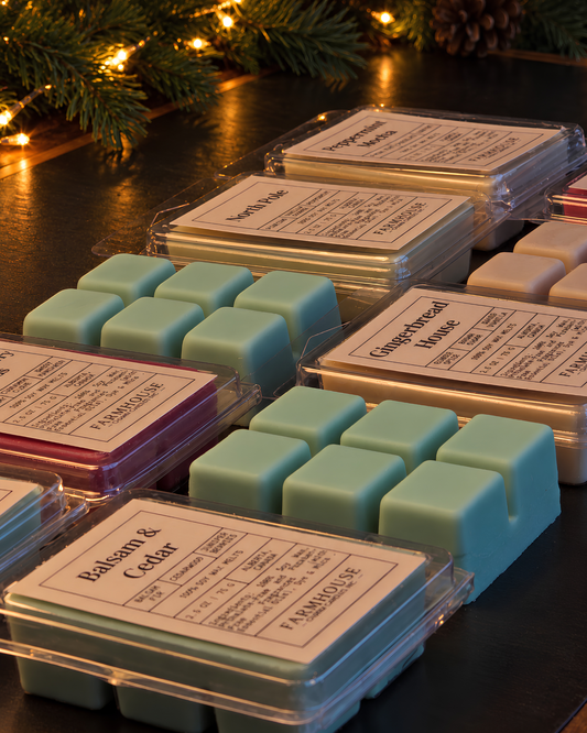 Several wax melts in clear plastic clamshell tins arranged on a dark tabletop, with pine branches and warm string lights in the background. The front-most tin shows sage green wax cubes and a label reading Home for Christmas Collection by Farmhouse Charm Candles