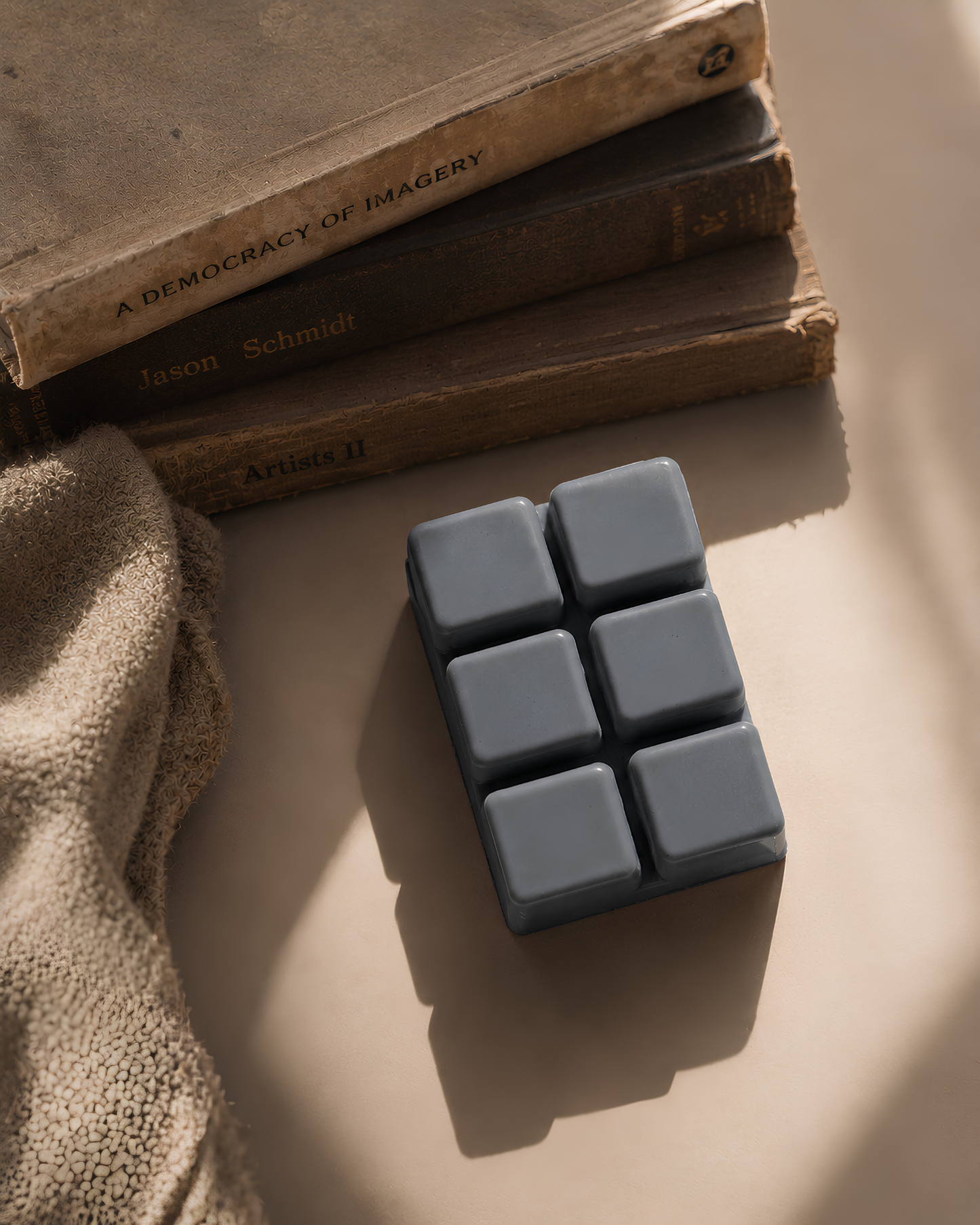 A rectangular bar of dark gray soap with six square sections, styled on a beige surface next to a textured beige fabric and a stack of vintage books. Farmhouse Charm Candles