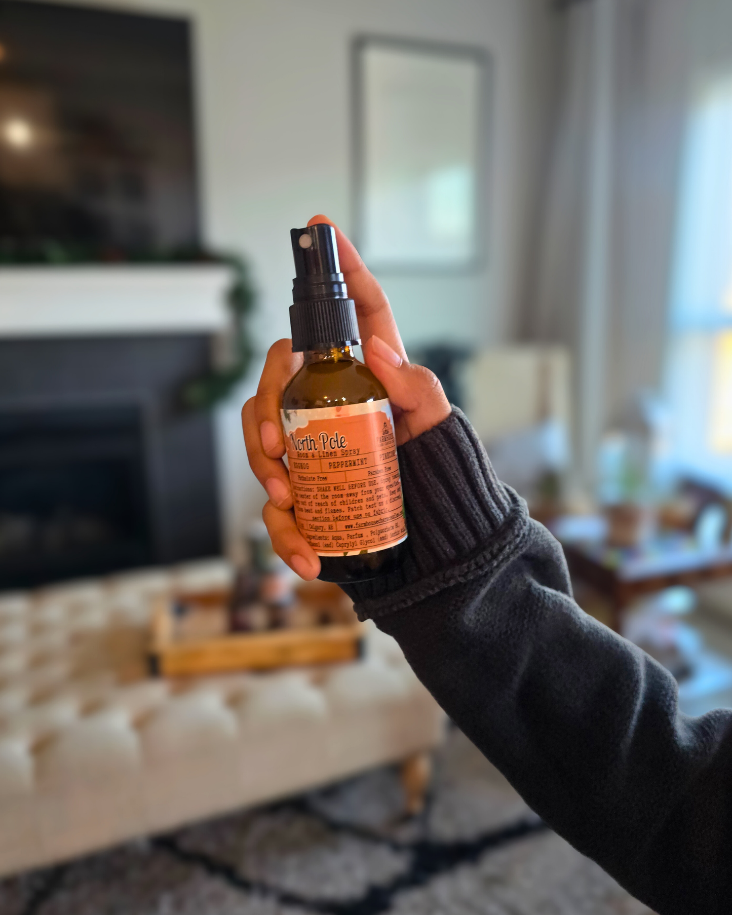 Hand holding a bottle of North Pole Room & Linen Spray capturing the cozy Christmas scent of eggnog, peppermint, and pinecone in a living room setting.