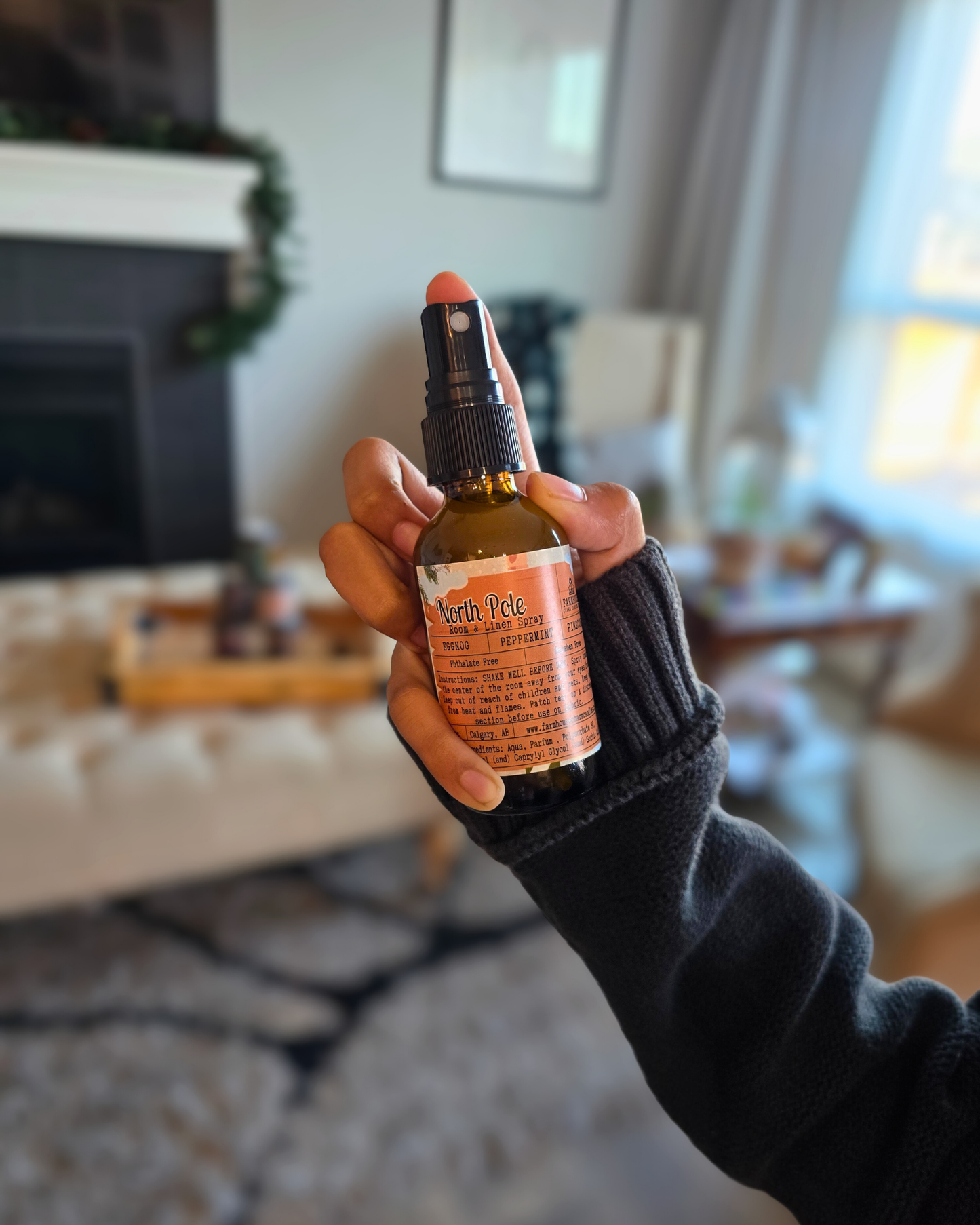 Hand holding a bottle of North Pole Room & Linen Spray capturing the cozy Christmas scent of eggnog, peppermint, and pinecone in a living room setting.