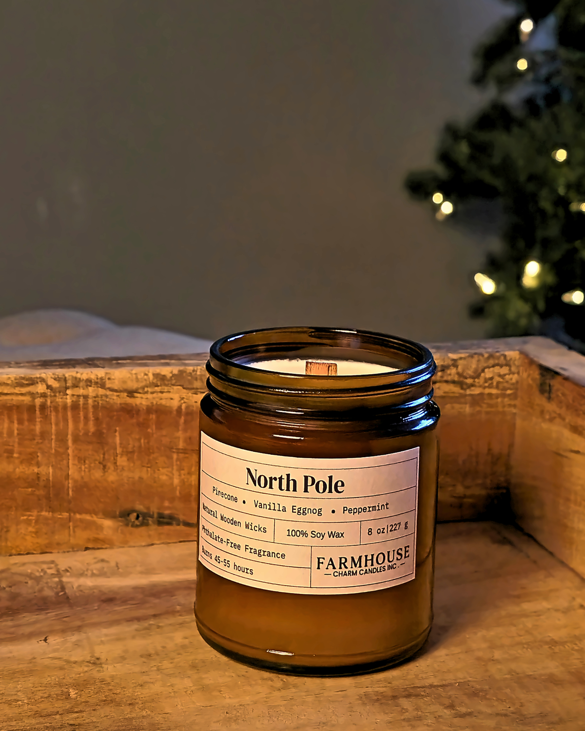 A Farmhouse Charm Candles 'North Pole' scented soy wax candle in an amber glass jar, set on a rustic wooden surface with a blurred Christmas tree in the background.