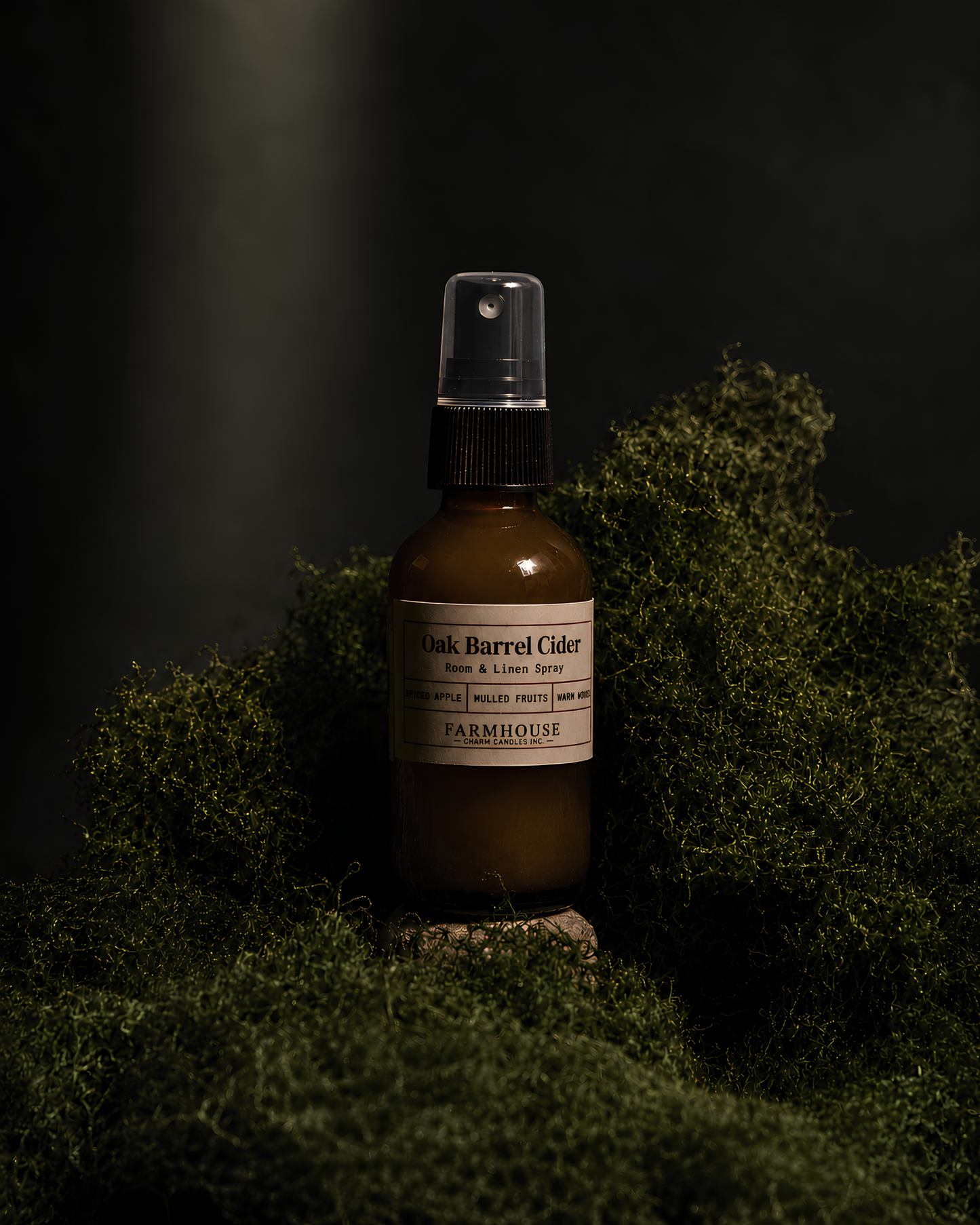 A bottle of Oak Barrel Cider Aromas & Linen Spray by Farmhouse Charm Candles, set in a dark, moody environment surrounded by green moss.