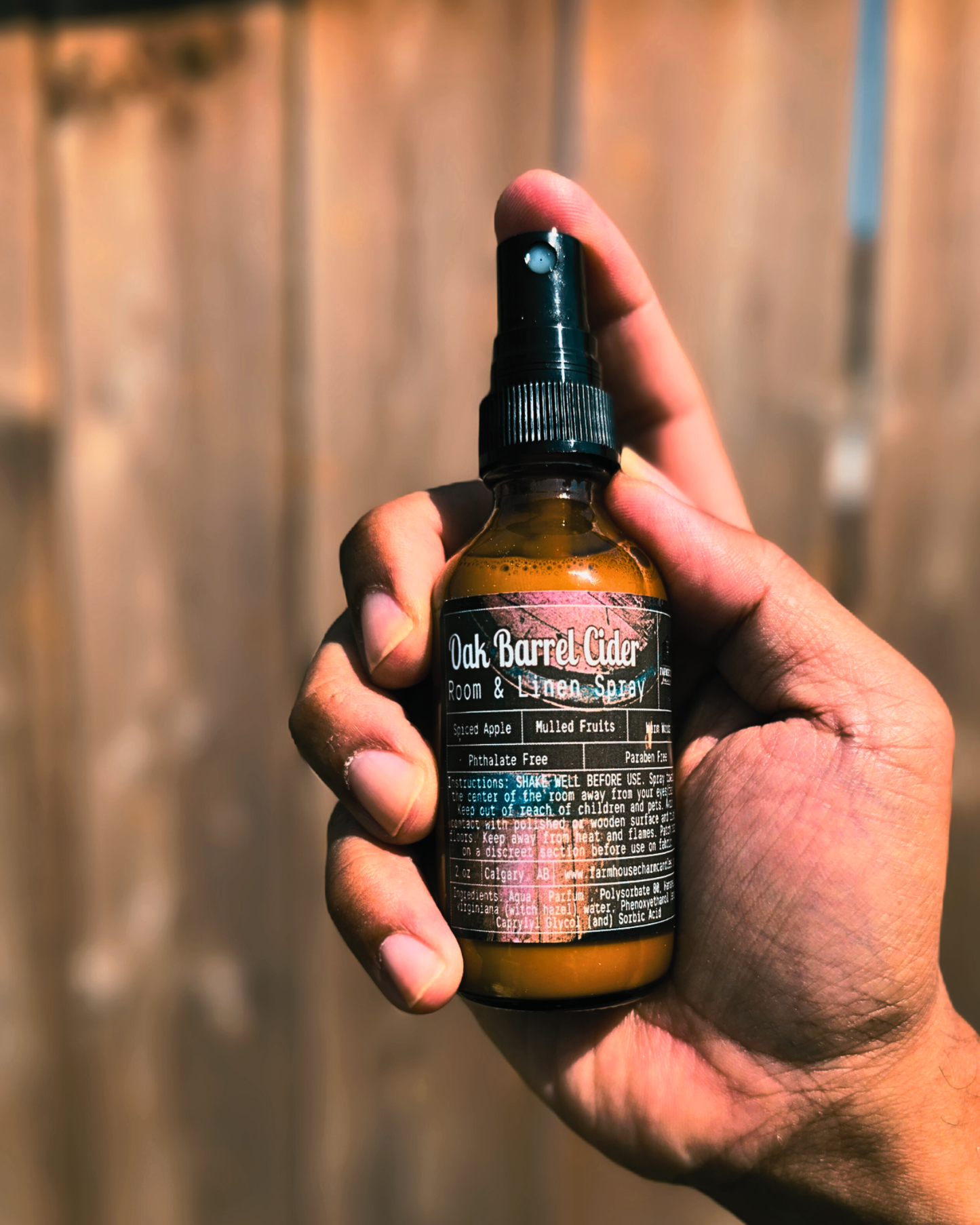 Hand holding a bottle of 'Oak Barrel Cider' Room and Linen Spray by Farmhouse Charm Candles with a fall themed background