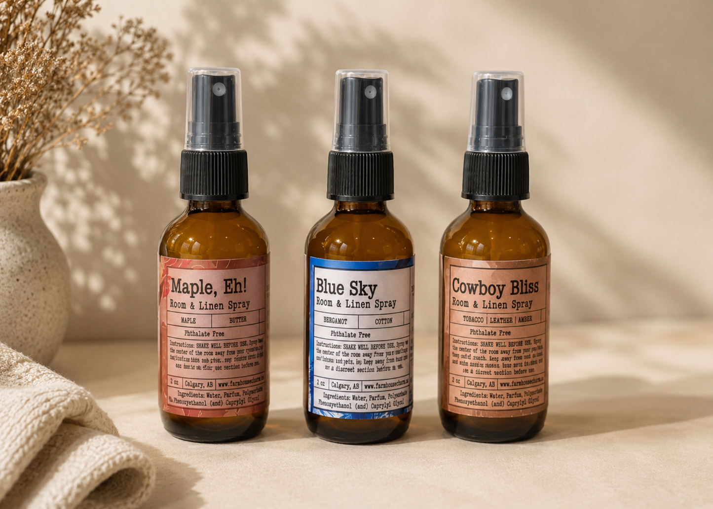 Oh Canada Room and Linen Spray Bundle — Maple Eh, Blue Sky and Cowboy Bliss amber glass spray bottles with black pumps on a warm beige surface beside dried pampas grass and a linen cloth — Farmhouse Charm Candles