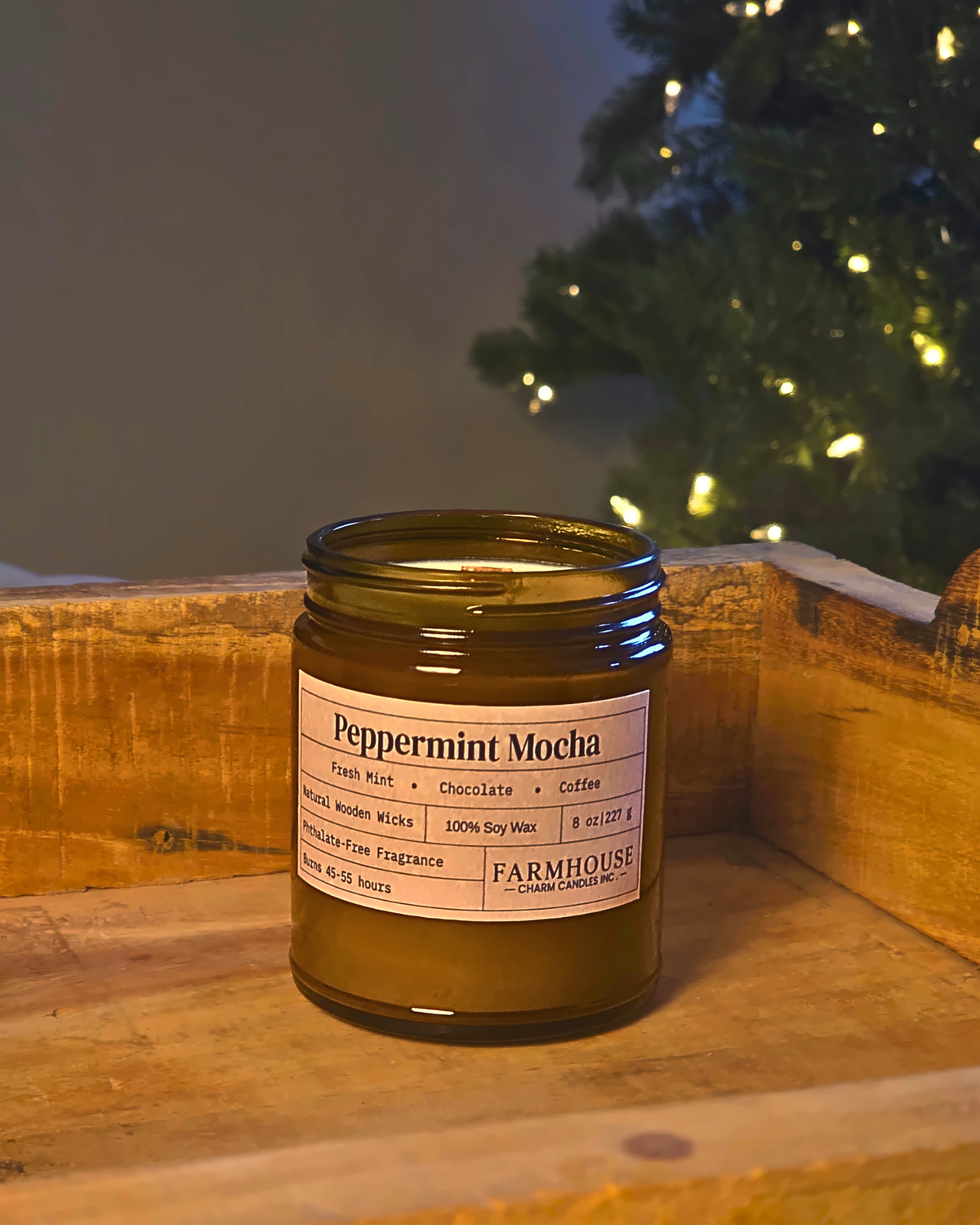 A Farmhouse Charm Candles 'Peppermint Mocha' scented candle in an amber glass jar, set on a rustic wooden crate with a blurred Christmas tree in the background.