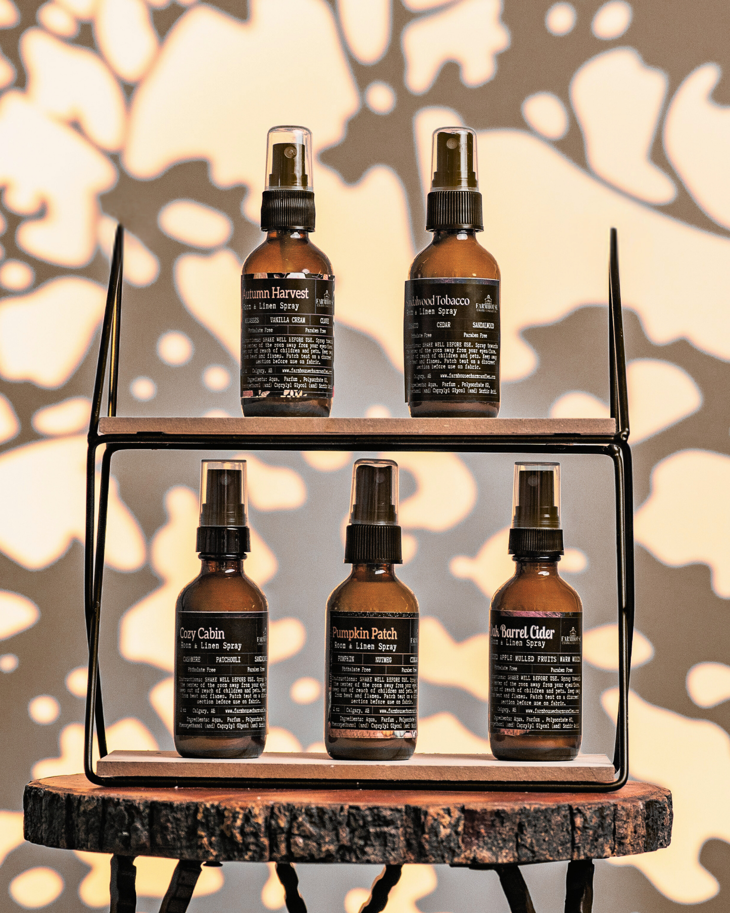 Set of five brown room & linen spray bottles by Farmhouse Charm Candles with labels on a wooden shelf against a leafy background