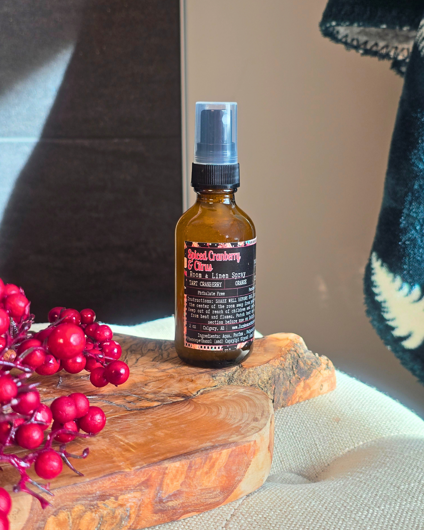 Amber glass bottle of Spiced Cranberry & Citrus Room and Linen Spray placed on a rustic wooden surface with holiday accents — evokes a warm, cozy Christmas scent of cranberry, orange, and cinnamon.