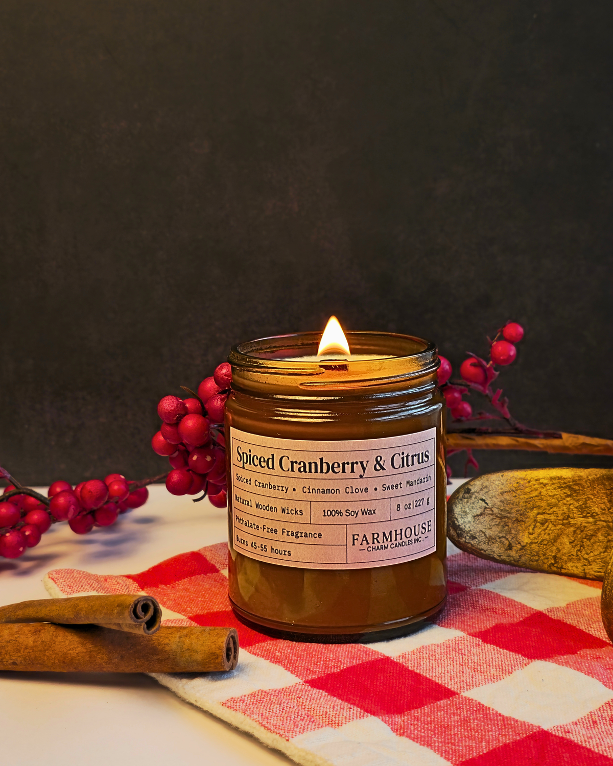 A lit candle in a clear glass jar with a white label reading 'Spiced Cranberry & Citrus' by Farmhouse Charm Candles styled on a red-and-white check cloth with cinnamon sticks, red berries, and a stone against a dark background.