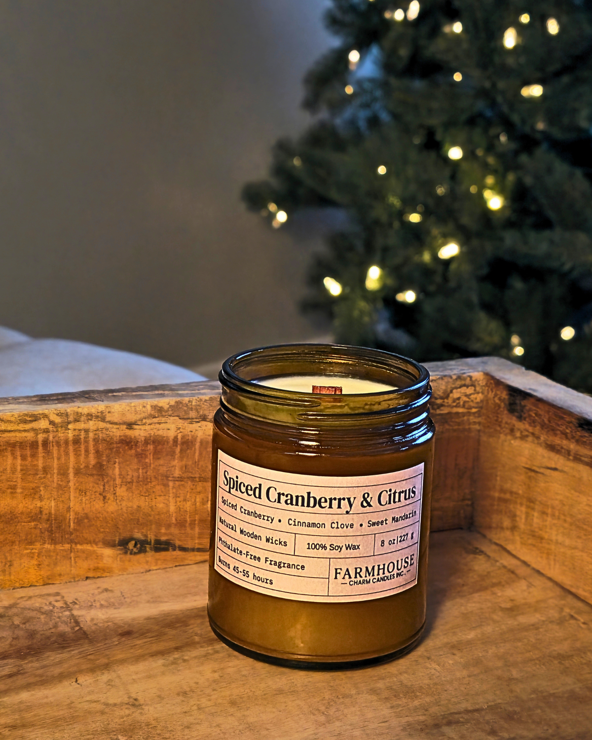 A Farmhouse Charm Candles jar candle in an amber glass jar with a white label reading 'Spiced Cranberry & Citrus,' placed on a wooden surface with a blurred Christmas tree in the background.
