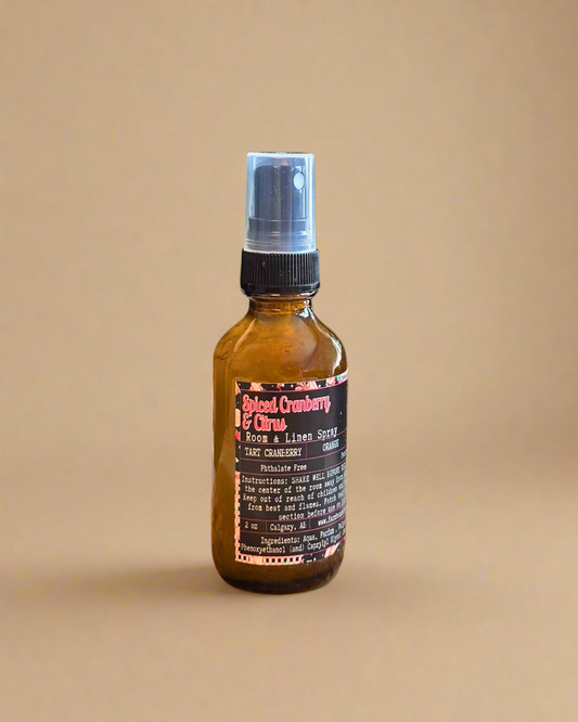 Amber glass bottle of Spiced Cranberry & Citrus Room and Linen Spray placed on a rustic wooden surface with holiday accents — evokes a warm, cozy Christmas scent of cranberry, orange, and cinnamon.