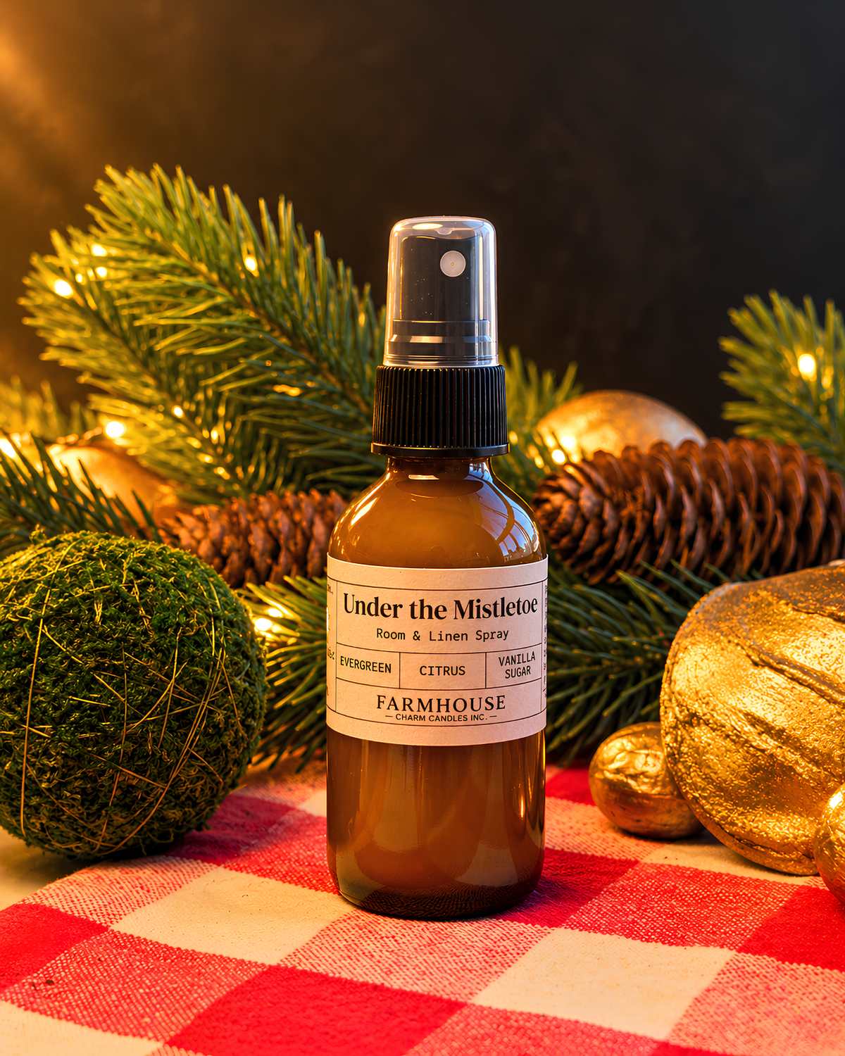 A small amber spray bottle labeled 'Under the Mistletoe' by Farmhouse Charm Candles stands on a red-and-white checkered cloth, surrounded by pine branches, pinecones, and gold holiday ornaments with warm string lights in the background.