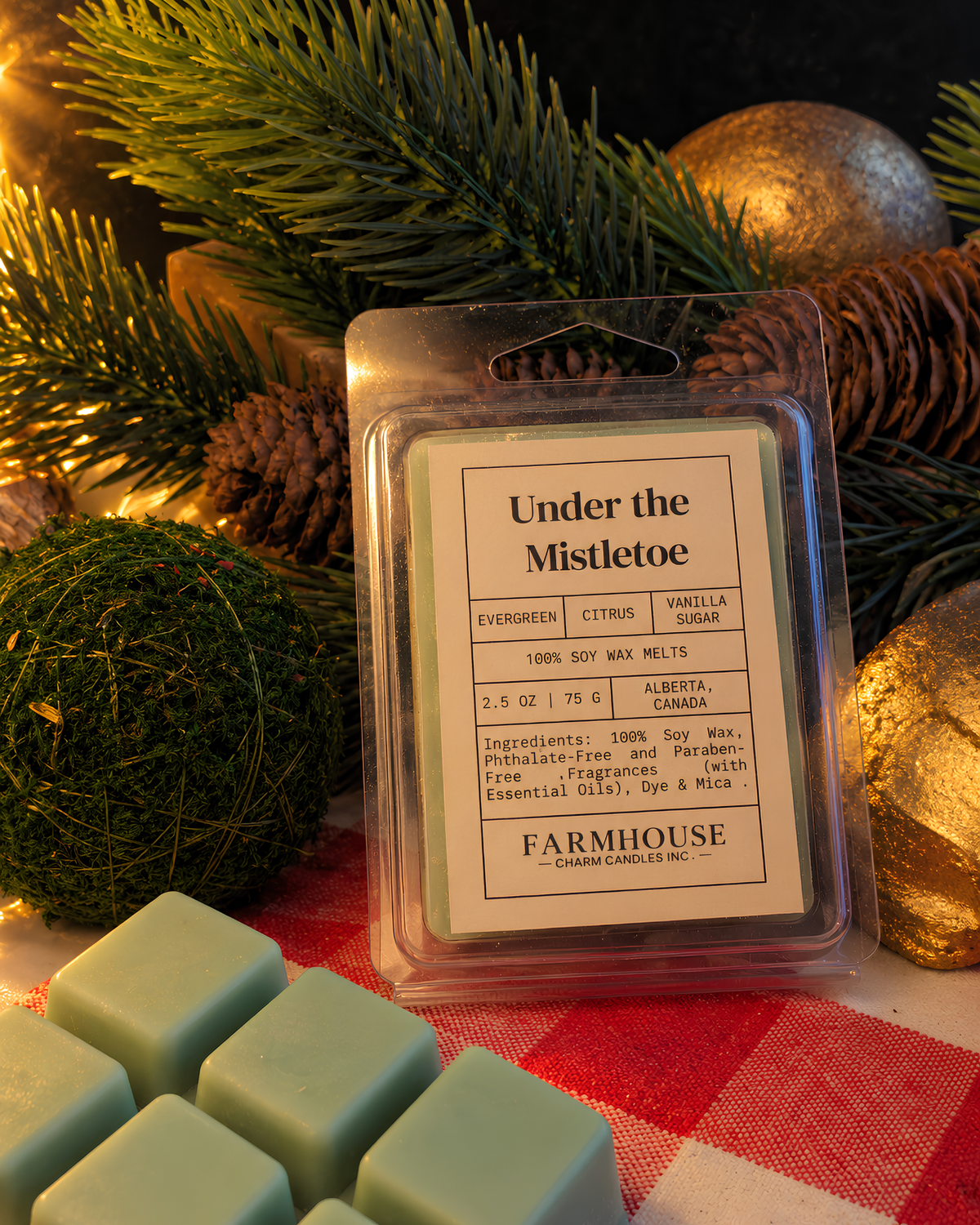 A packaged Farmhouse Charm Candles Inc. 'Under the Mistletoe' citrus vanilla soy wax melt sits on a red-and-white checkered blanket with pine branches, pinecones, and green moss balls around it. Several pale mint wax cubes are visible in the foreground.