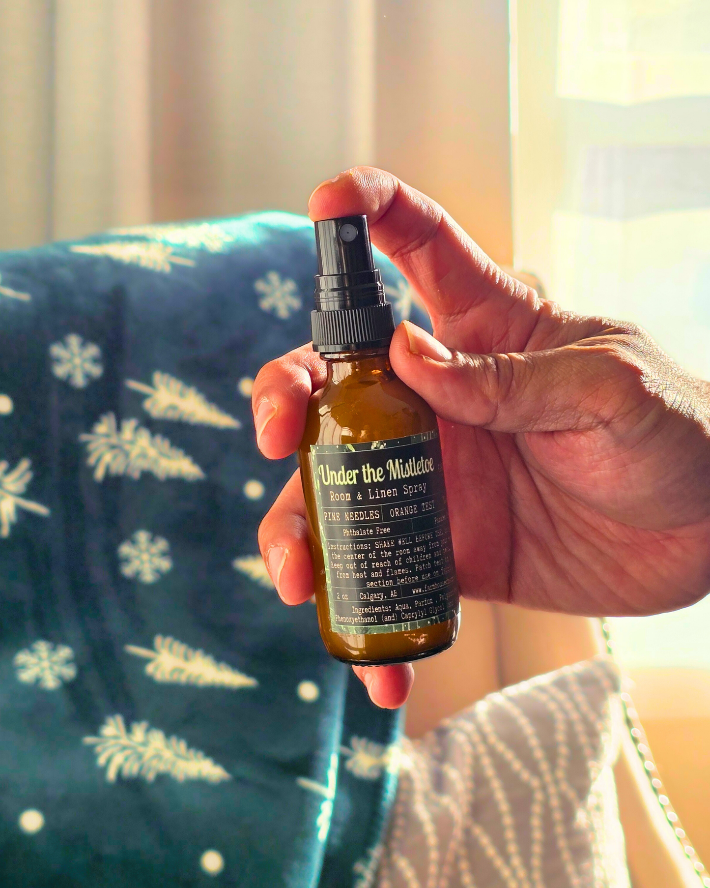 Hand holding a Brown spray bottle with label ' Under the Mistletoe by Farmhouse charm Candles on cozy living room