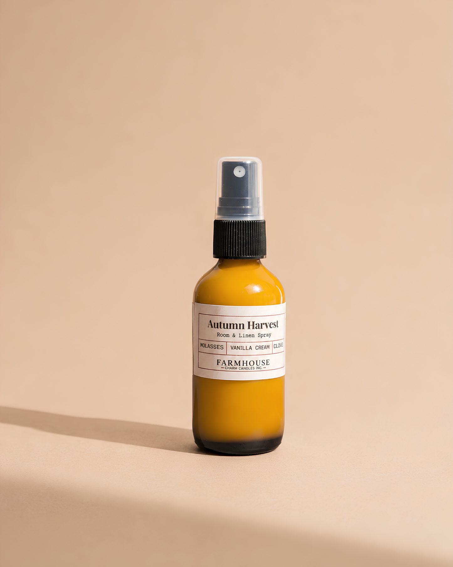 A small amber glass spray bottle with a white label reading 'Autumn Harvest' by Farmhouse Charm Candles on a warm beige background.
