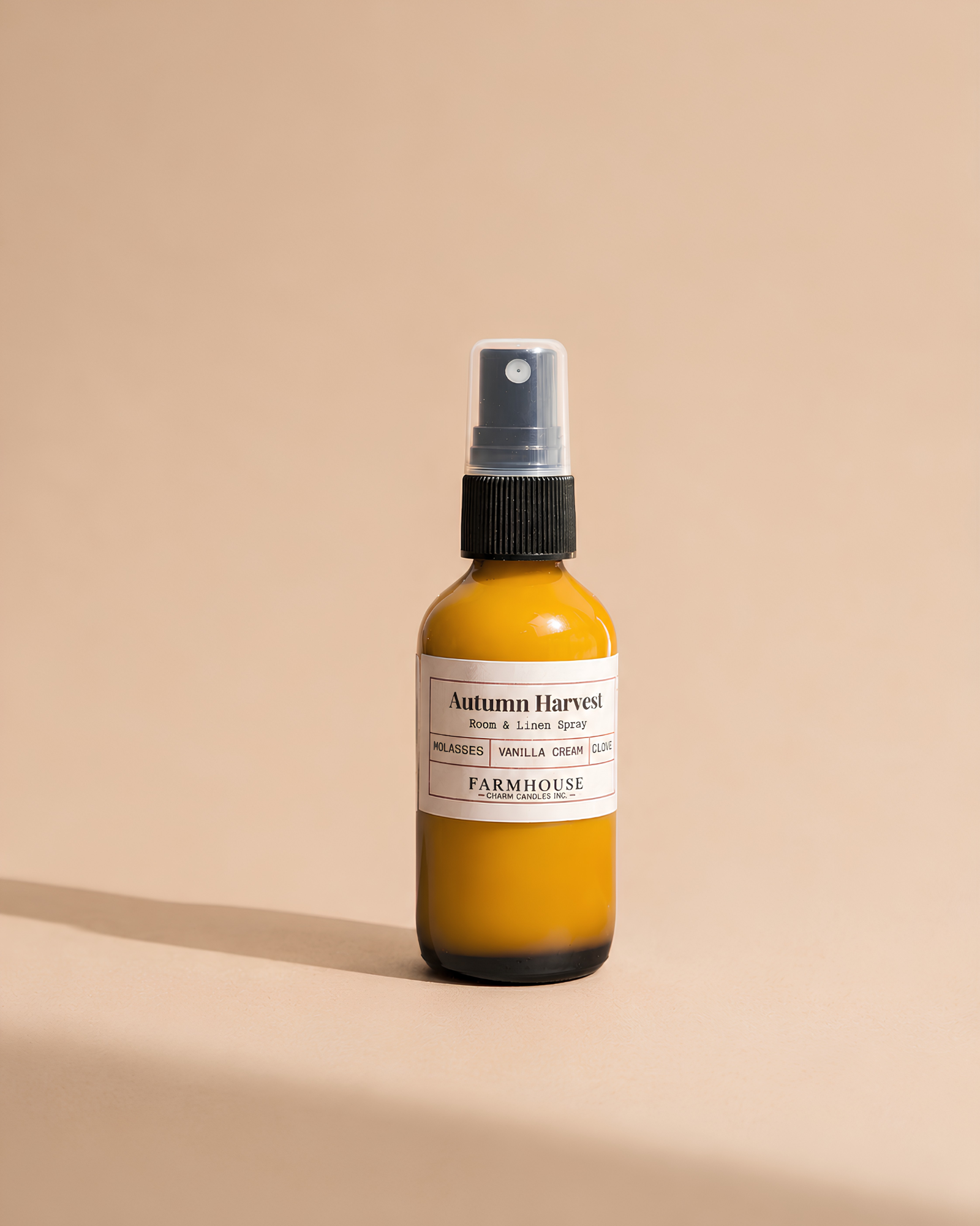 A small amber glass spray bottle with a white label reading 'Autumn Harvest' by Farmhouse Charm Candles on a warm beige background.