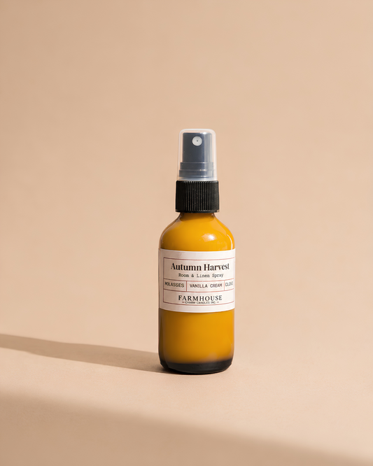 A small amber glass spray bottle with a white label reading 'Autumn Harvest' by Farmhouse Charm Candles on a warm beige background.