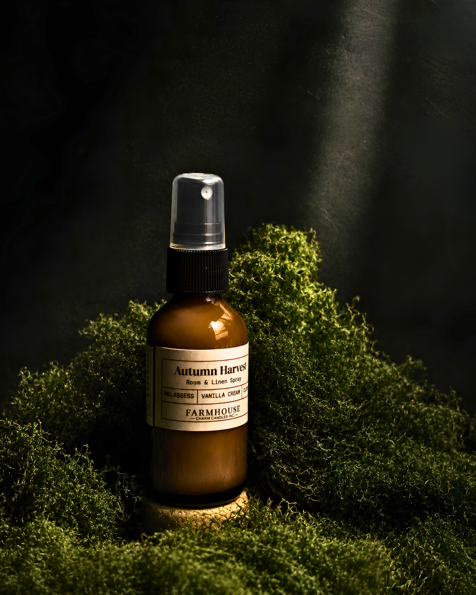 An amber glass spray bottle labeled 'Autumn Harvest' by Farmhouse Charm Candles , placed on a bed of green moss against a dark, moody background with dramatic light beams.