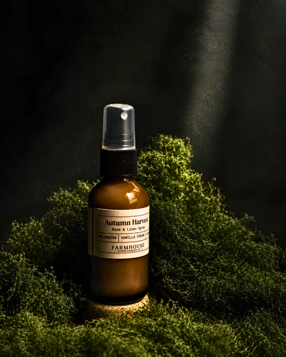 An amber glass spray bottle labeled 'Autumn Harvest' by Farmhouse Charm Candles , placed on a bed of green moss against a dark, moody background with dramatic light beams.