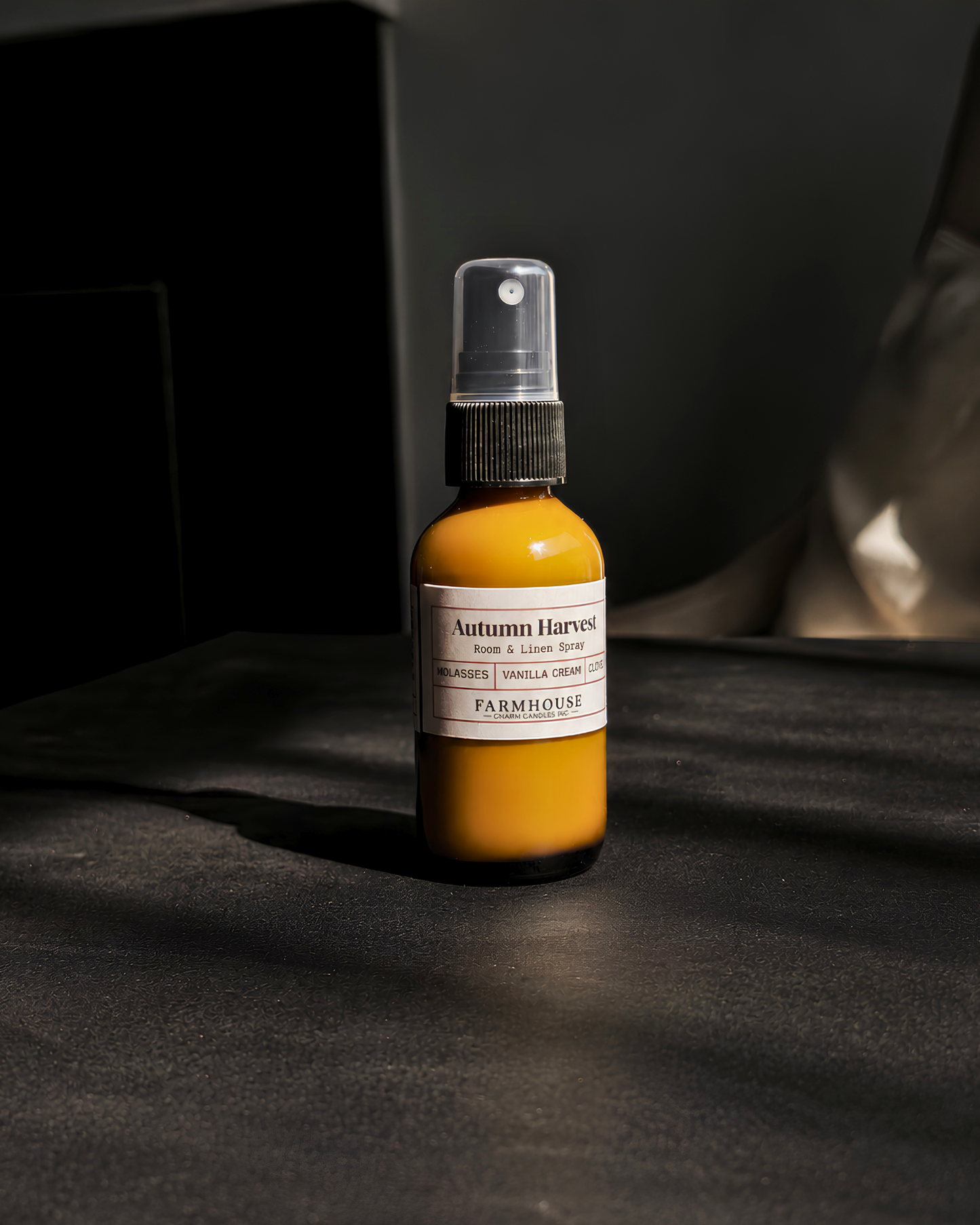 A small amber spray bottle with a white label reading 'Autumn Harvest' by Farmhouse Charm Candles  stands on a dark surface in dramatic side lighting against a dark background.