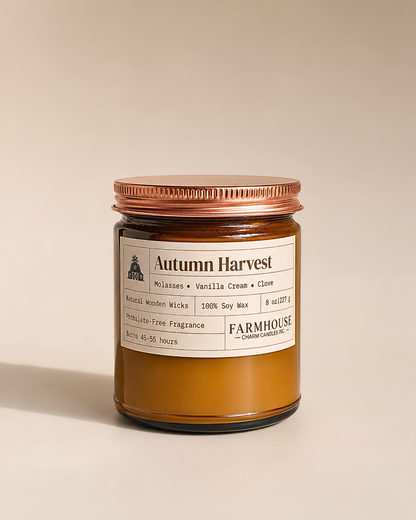A Farmhouse Candle Company 'Autumn Harvest' scented candle in a amber-colored glass jar and a copper metal lid, set against a solid beige background.