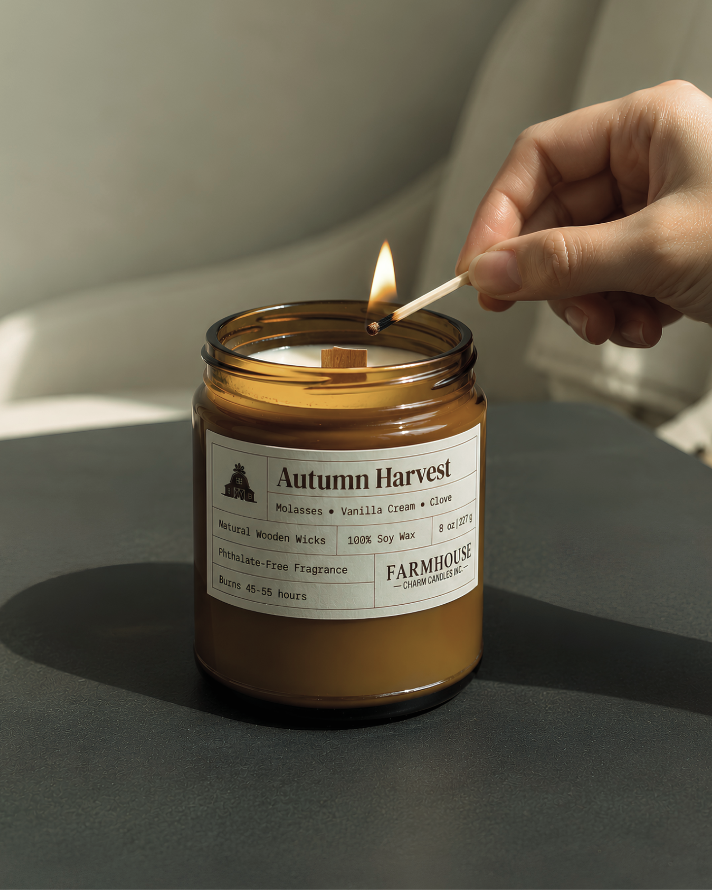 An amber glass jar candle labeled 'Autumn Harvest' by Farmhouse Charm Candles  is lit with a wooden match held by a person's hand, set on a dark surface against a soft neutral background.