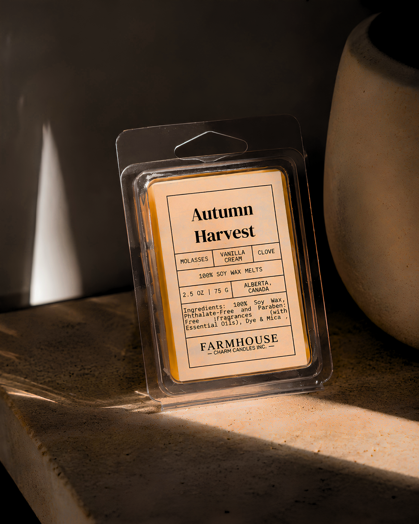 A packaged wax melt labeled 'Autumn Harvest' by Farmhouse Charm Candles package on a stone surface with warm sunlight and shadows.