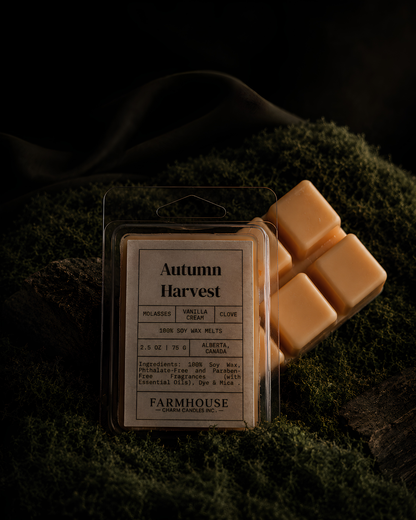 A packaged wax melt product labeled 'Autumn Harvest' sits on dark green moss with several pale peach wax cubes beside it. The scene is styled in low light with a dark fabric backdrop, and the visible brand at the bottom of the label is Farmhouse Charm  Candles