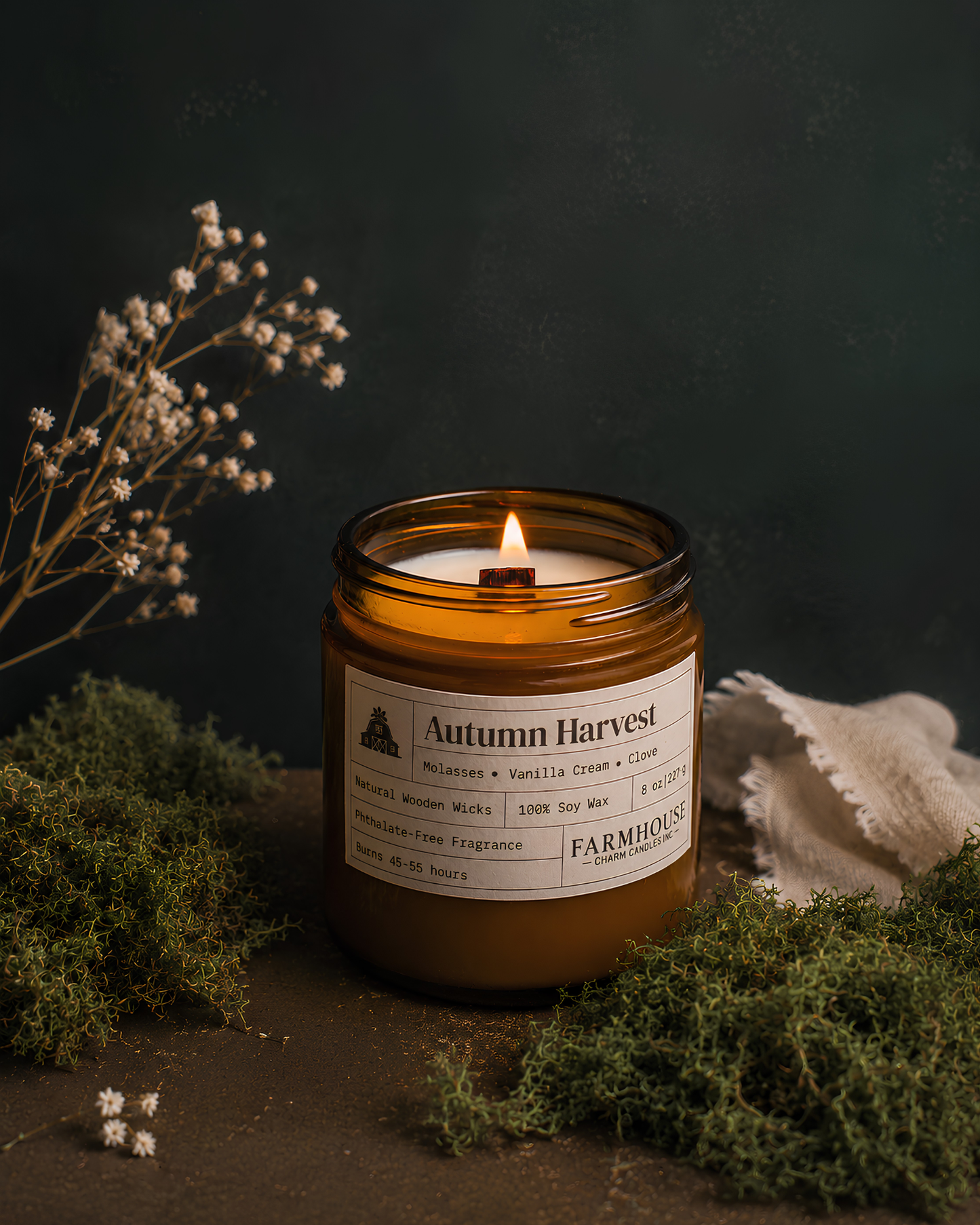 A lit amber glass jar candle labeled Autumn Harvest by Farmhouse Charm Candles, styled on a dark surface with dried white flowers, green moss, and a piece of textured white fabric against a dark green background.