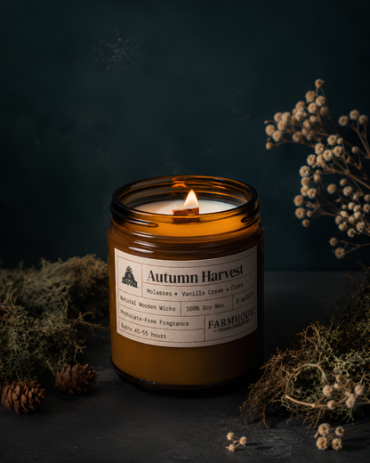 A lit amber glass jar candle labeled 'Autumn Harvest' by Farmhouse Charm Candles, styled with dried botanicals, pinecones, and moss on a dark textured surface.