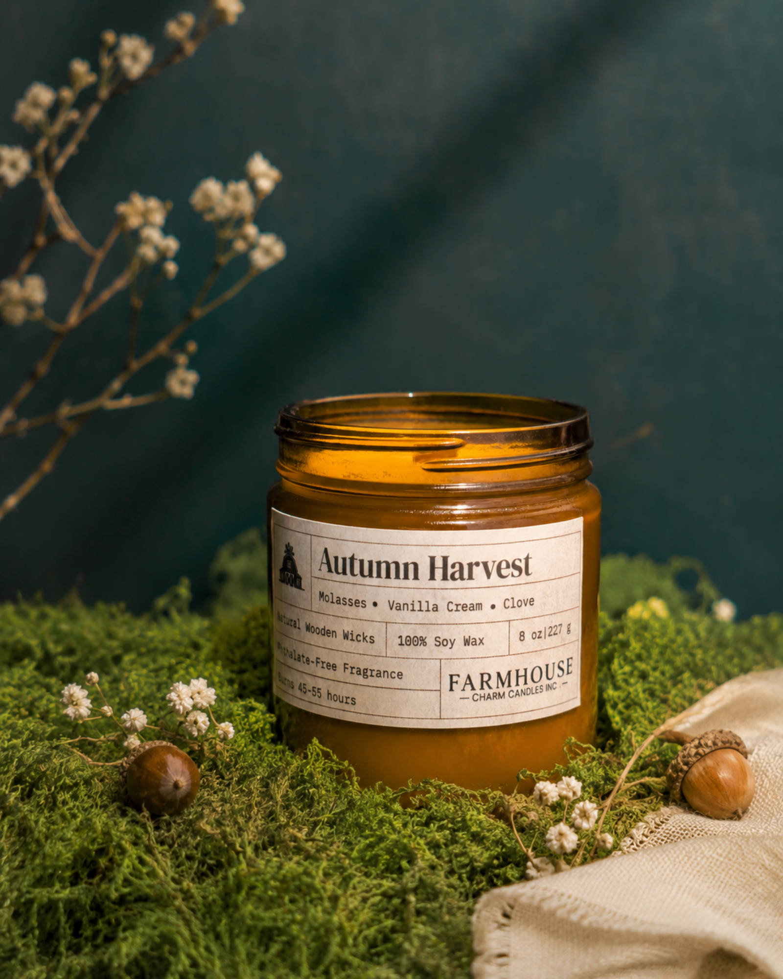 An amber glass jar candle labeled 'Autumn Harvest' by Farmhouse Charm Candles., styled on moss with small white flowers, acorns, and a beige cloth against a dark teal background.