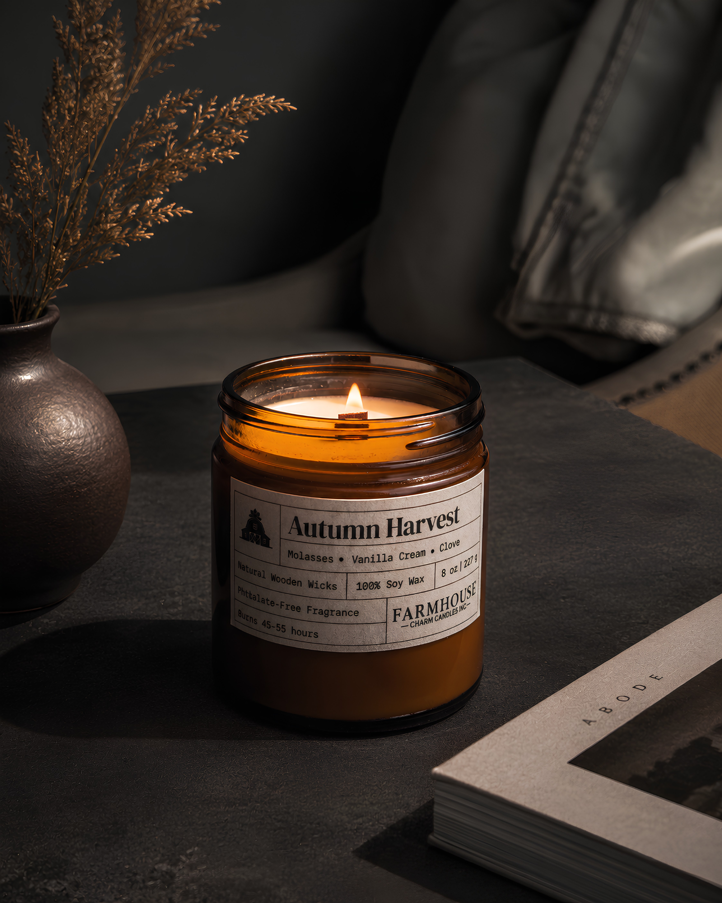 A lit amber glass jar candle labeled 'Autumn Harvest' by Farmhouse Candle Co., placed on a dark tabletop beside a vase with dried stems and an open book in a moody indoor setting.