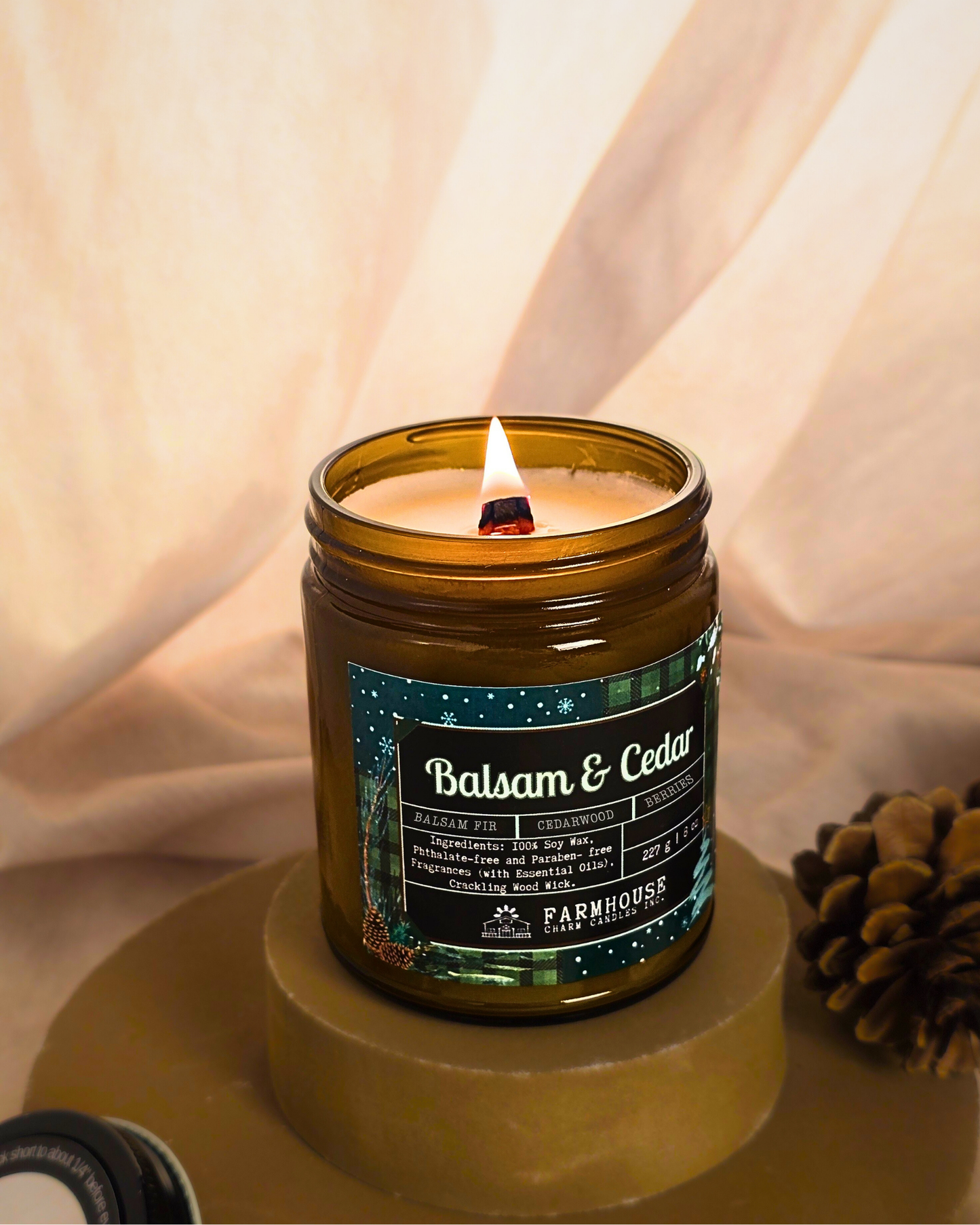 Candle in a glass jar labeled 'Balsam & Cedar' by Farmhouse Charm Candles on a wooden surface with pinecones.