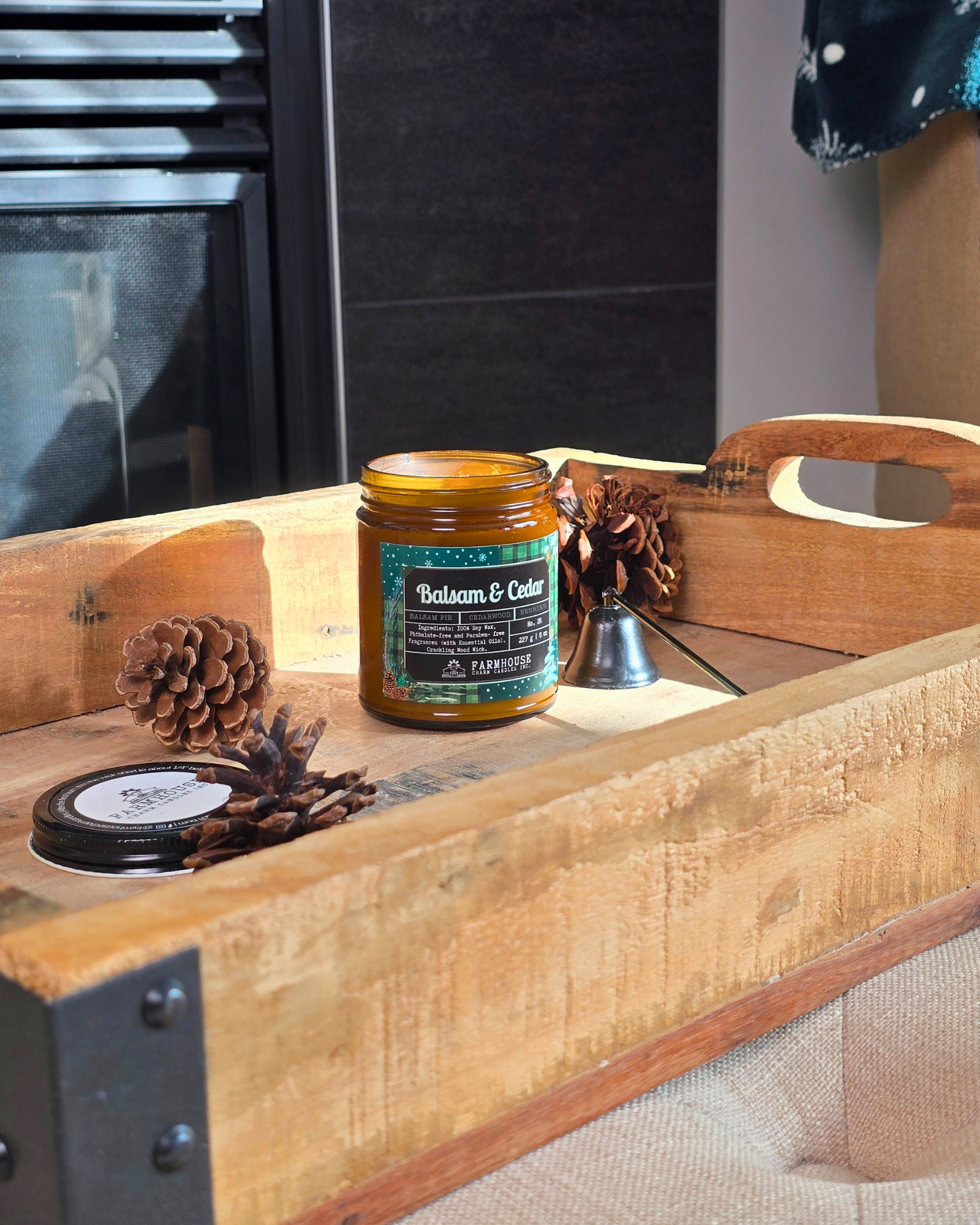 Candle in a glass jar labeled 'Balsam & Cedar' by Farmhouse Charm Candles on a wooden surface with pinecones.