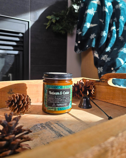 Candle in a glass jar labeled 'Balsam & Cedar' by Farmhouse Charm Candles on a wooden surface with pinecones.